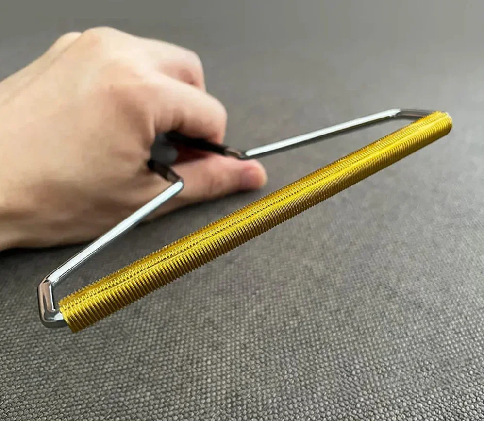 Hand holding metal pet hair remover tool with gold bristles on gray fabric background